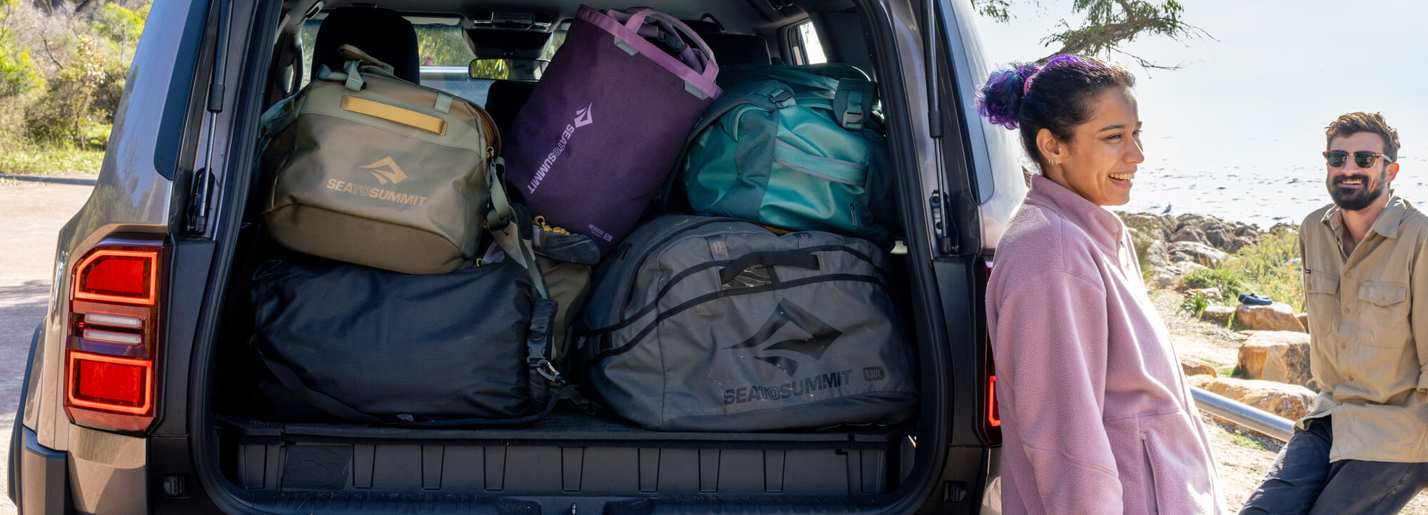 A woman and a man stand beside the open trunk of a vehicle loaded with multiple Sea to Summit duffle and waterproof tote packs in olive, purple, teal, and gray, next to a beach.